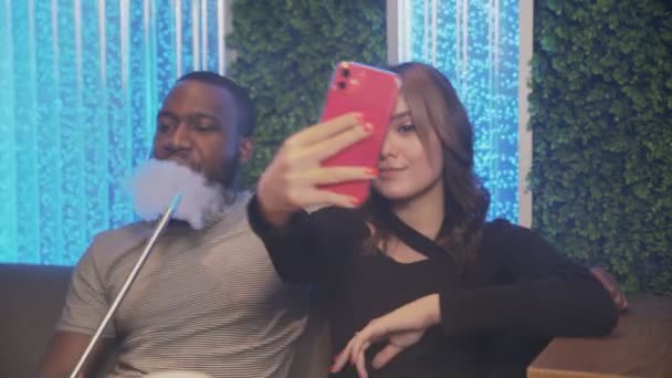 Young woman takes selfie on phone, while her african boyfriend smokes hookah - 映像、動画