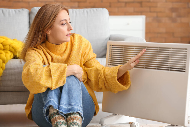 Mature woman near electric heater at home. Concept of heating season - Photo, Image