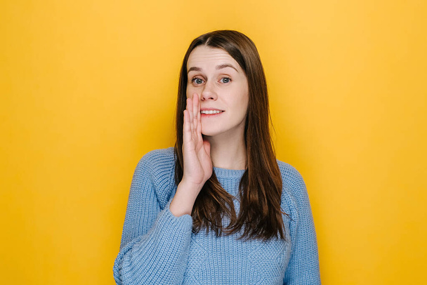 Serious young woman look at camera tell secret information, millennial cute brunette girl make loudspeaker share private sale promotion, dressed in blue sweater, isolated on yellow studio background - Photo, Image