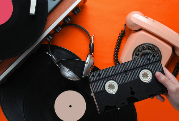 Female hand holds video cassette. 80s style. Vinyl player, headphones, rotary phone on orange  background. Top view, flat lay - Photo, Image