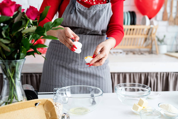 Ημέρα του Αγίου Βαλεντίνου. Γυναίκα με κόκκινο φόρεμα και γκρι ποδιά κάνοντας Valentine μαγείρεμα στην κουζίνα - Φωτογραφία, εικόνα