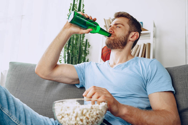 Young bearded man sitting on couch with beer and popcorn while watching movie - Foto, immagini