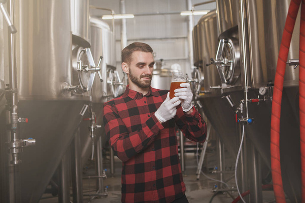 Όμορφος επαγγελματίας ζυθοποιός εξετάζει πρόσφατα παρασκευασμένη μπύρα σε ένα ποτήρι στο μικροζυθοποιείο του - Φωτογραφία, εικόνα