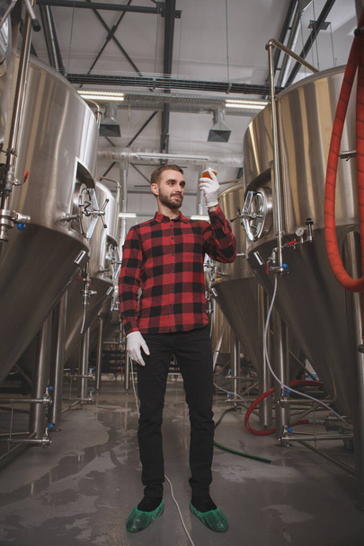 Küçük bira fabrikasında bira içen yakışıklı bir adamın düşük açılı dikey çekimi. Metal bira tanklarının arasında duruyor. - Fotoğraf, Görsel