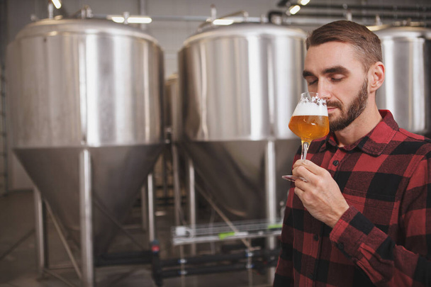 Aantrekkelijke mannelijke brouwer die aromatisch vers gebrouwen bier ruikt in de microbrouwerij, kopieerruimte - Foto, afbeelding