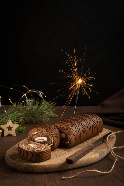 Chocolate sponge roll with white cream on wooden cutting board with knife and firework sparkler Bengal light on dark table against black background at winter holiday with fir tree and towel. Vertical - Fotó, kép