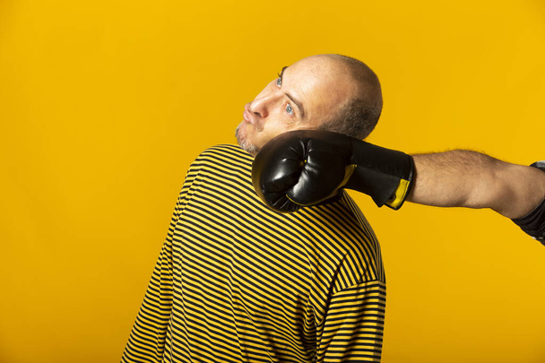 Mann mittleren Alters bekommt Faustschläge in den Kiefer. Hand eines Mannes mit Boxhandschuhen schlägt anderen Mann vor gelbem Hintergrund ins Gesicht - Foto, Bild
