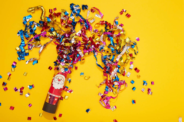 Colorful bright confetti and streamers with red party cracker on yellow background, top view. Copy space for text. Santa Claus on the clapperboard. Mood of Christmas and happy new year. - Photo, Image