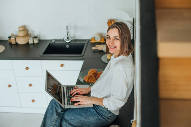 Fiatal mosolygós barna nő alkalmi ruhák ül az asztalon a konyhában segítségével laptop a munka vagy üzenetküldés. - Fotó, kép