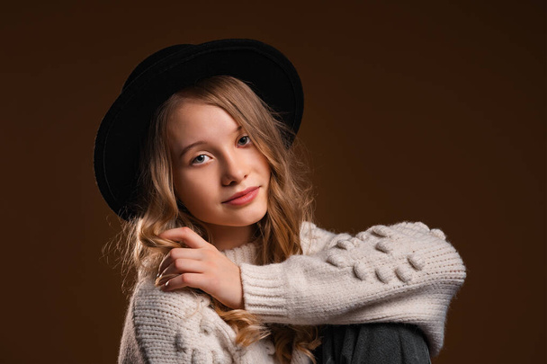 Charming blonde teen girl in black hat posing in studio on brown background. Concept of beauty and elegance - Fotografie, Obrázek