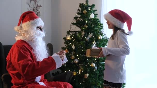 Санта Клаус дарит милой белой девушке рождественский или новогодний подарок дома. Счастливый ребенок в санта-шляпе с подарком - Кадры, видео