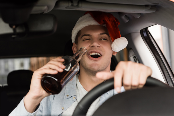 Betrunkener, aufgeregter Mann mit Weihnachtsmütze, der Auto fährt und eine Flasche Whiskey im verschwommenen Vordergrund hält - Foto, Bild
