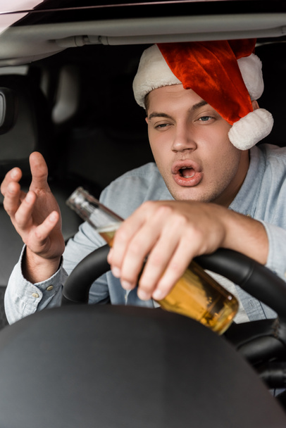 borracho hombre en santa hat celebración botella de alcohol y gesto mientras conduce coche en primer plano borrosa - Foto, Imagen