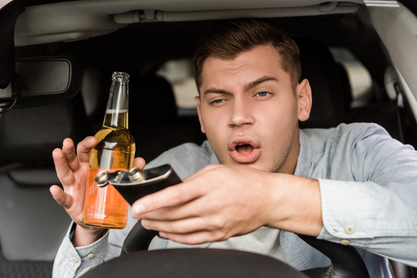 пьяный мужчина держит бутылку виски и фляжку, сидя в машине - Фото, изображение
