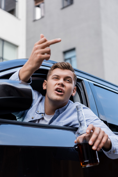 Betrunkener, aggressiver Mann mit Whiskeyflasche zeigt Mittelfinger beim Blick aus dem Autofenster, verschwommener Vordergrund - Foto, Bild
