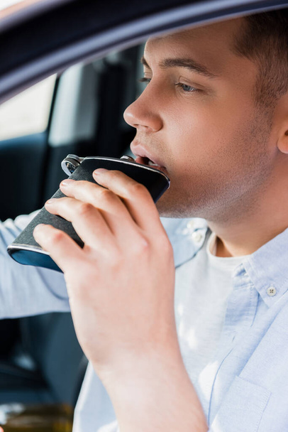 young man drinking alcohol from flask while driving car, blurred foreground - Foto, Imagen