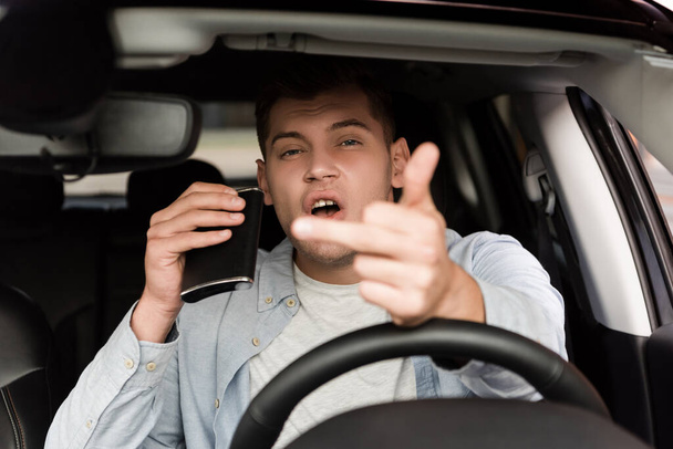 drunk man holding flask with alcohol and showing middle finger in car, blurred foreground - Photo, Image