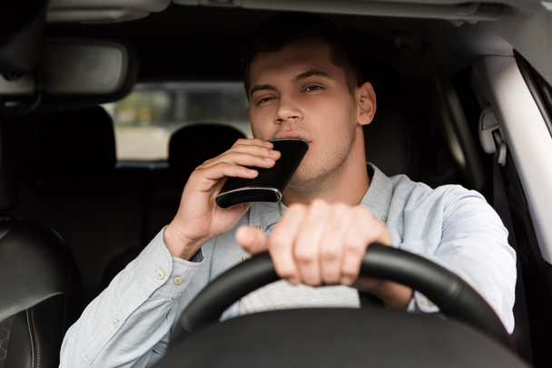 mladý muž pije alkohol z baňky při řízení auta, rozmazané popředí - Fotografie, Obrázek