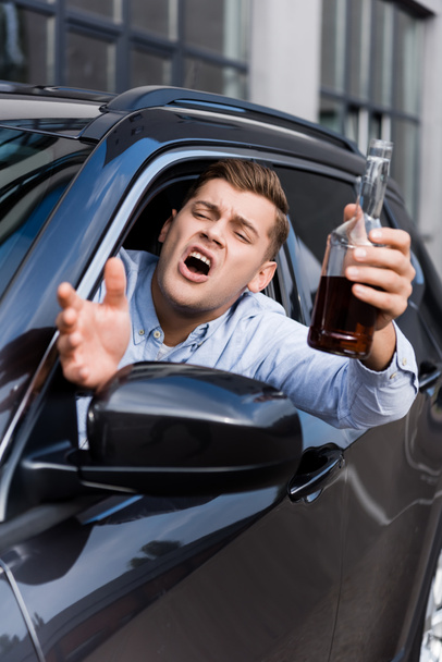 borracho, hombre enojado con botella de alcohol haciendo gestos y gritando mientras mira por la ventana del coche, borroso primer plano - Foto, Imagen