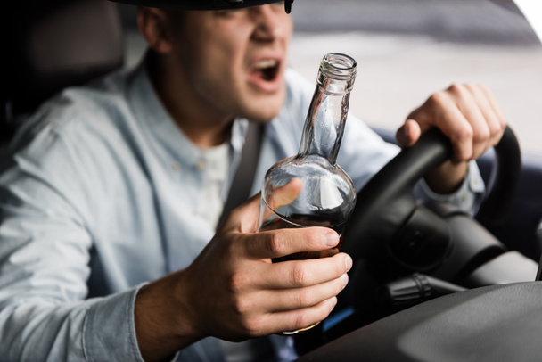 vista recortada de agresivo, borracho hombre con botella de whisky gritando mientras conduce coche - Foto, Imagen