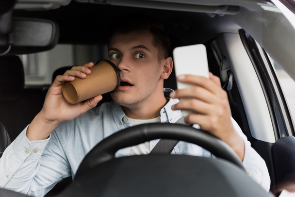 surprised man drinking coffee to go and using smartphone at steering wheel in car, blurred foreground - Foto, imagen