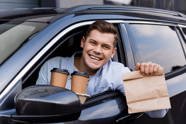 conductor alegre sosteniendo café para ir y bolsa de papel mientras sonríe a la cámara - Foto, Imagen