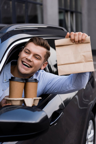 szczęśliwy człowiek gospodarstwa jednorazowy kubek i torba papierowa podczas siedzenia w samochodzie - Zdjęcie, obraz
