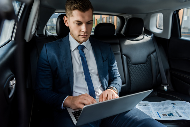 Young businessman using laptop near newspaper on black seat of auto - Photo, Image