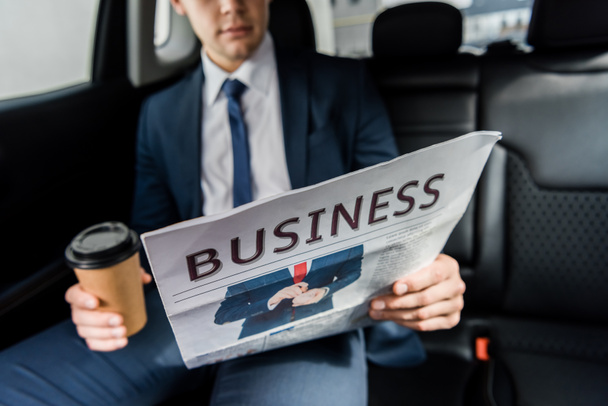 Vista recortada del periódico en la mano del hombre de negocios con café para ir en coche sobre fondo borroso - Foto, Imagen