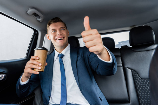 Positive businessman holding takeaway coffee and showing like gesture on blurred foreground in auto - Photo, Image