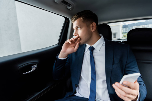 Businessman looking at window and holding smartphone on blurred foreground in car - Zdjęcie, obraz