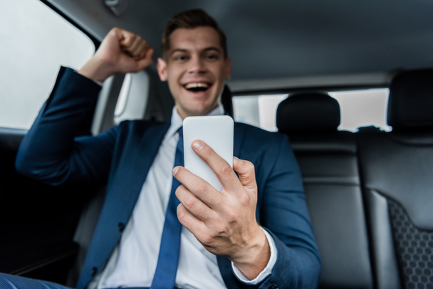 Smartphone in hand of cheerful businessman showing yeah gesture on blurred background in car - Foto, imagen