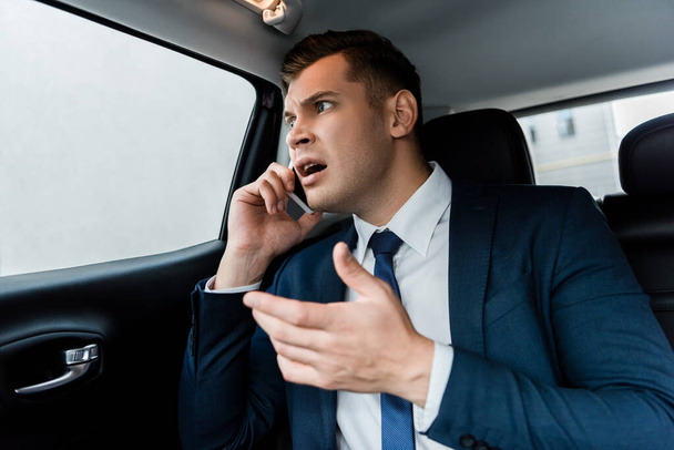 怒っているビジネスマンは車の後部座席でスマートフォンで話す - 写真・画像