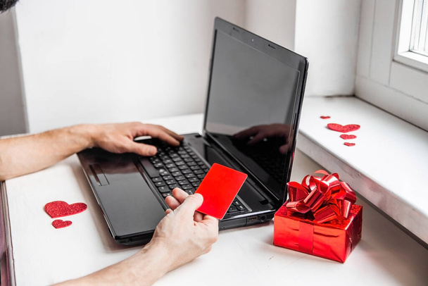 Valentines day online shopping concept. Close up of a mans hands hold a red mock up of credit card and gift near a laptop. - Photo, Image