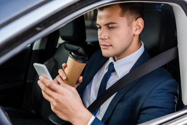 Young businessman using smartphone and holding coffee to go on driver seat in car - 写真・画像