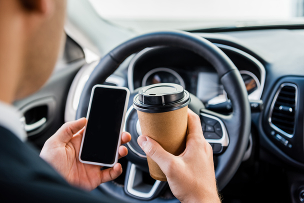 Cropped view of man holding smartphone with blank screen and takeaway coffee in car on blurred background - Photo, Image