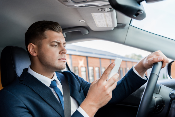 Concentrated businessman using smartphone while driving car - 写真・画像