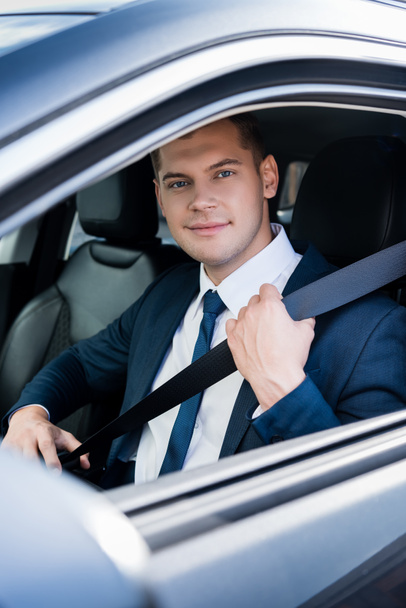 Young businessman looking at camera while holding seatbelt in auto - 写真・画像