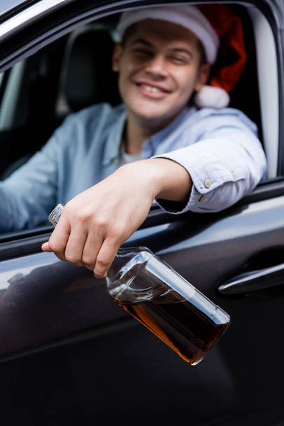 dronken man in santa hoed glimlachen en het houden van fles whisky in de auto op wazig achtergrond - Foto, afbeelding