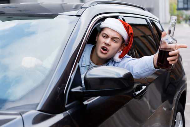 borracho, hombre enojado en sombrero de santa, con botella de whisky, mirando por la ventana mientras conduce el coche - Foto, Imagen