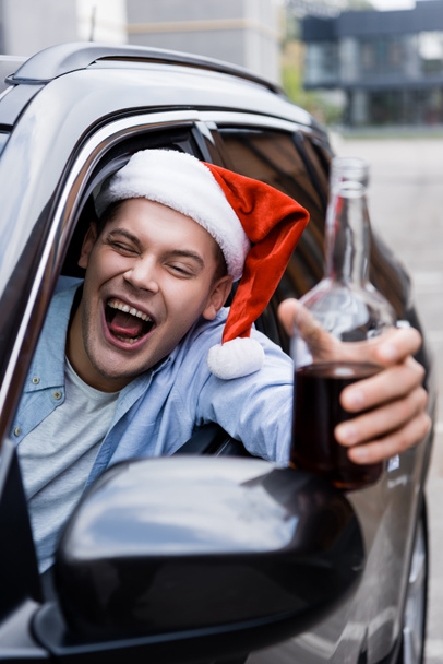 drunk man in santa hat grimacing and holding bottle of whiskey in car on blurred foreground - Photo, Image