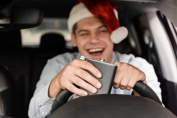 lachend, Betrunkener mit Weihnachtsmannmütze hält Flasche mit Alkohol während Autofahrt auf verschwommenem Hintergrund - Foto, Bild