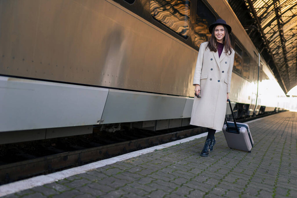 Chica positiva con maleta camina en la plataforma de la estación de tren cerca de vagón de tren - Foto, imagen