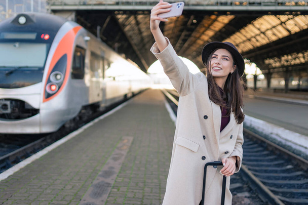 Tourist woman in joyful mood makes selfie while standing on the platform of the railway station - Foto, imagen