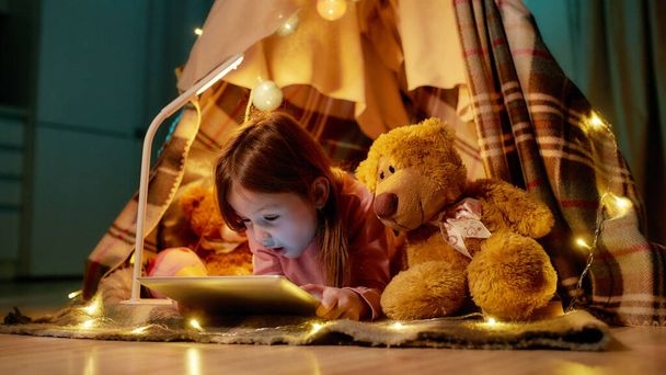 A cute small girl lying on a floor covered with a plaid barefoot in a self-made hut made of a plaid holding a tablet and watching videos with her teddybear and garlands all around the hut - Foto, Imagen