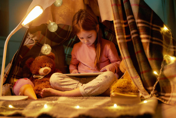 A cute little girl wearing pyjamas sitting on a floor cross-legged barefoot in a self-made hut made of a plaid watching cartoons on a tablet with her teddybear and fancy garlands around - Photo, image