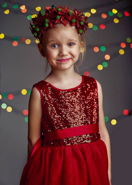 Χριστουγεννιάτικο πορτρέτο χαρούμενο νεαρό κορίτσι που φοράει κόκκινο φόρεμα με πούλιες και Χριστουγεννιάτικο στεφάνι. Ξανθά μαλλιά και μπλε μάτια ποζάρουν και παίζουν στο στούντιο σε γκρι φόντο με φωτισμό και boke φώτα. - Φωτογραφία, εικόνα