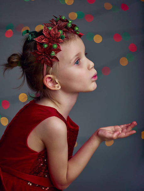 Christmas kiss. Kid girl wearing red sequin dress and Christmas wreath. Blond hair and blue eyes posing and fool around in studio on grey background with illumination and boke lights. - Photo, Image