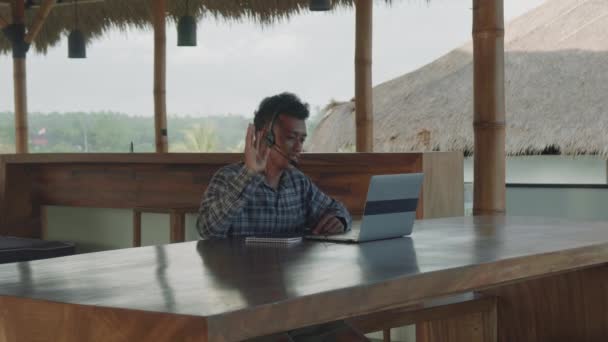 Mediana toma de hombre asiático joven con ropa casual y auriculares sentados en la mesa de madera en la terraza en la zona tropical, mirando a la pantalla del ordenador portátil y el cliente de consulta por enlace de vídeo - Metraje, vídeo