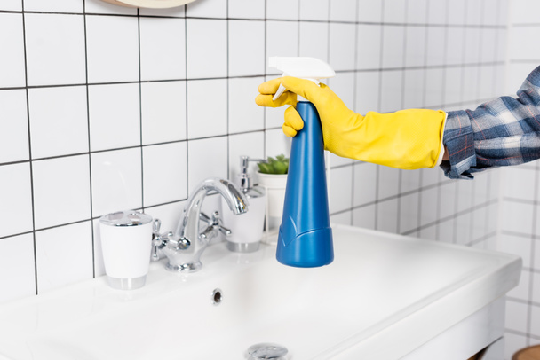 Widok kobiety w gumowej rękawicy trzymającej butelkę detergentu w pobliżu zlewu i płytek w łazience  - Zdjęcie, obraz